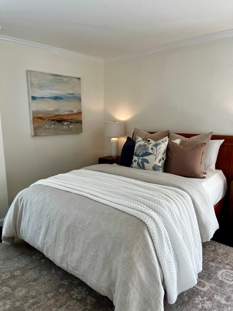 A cozy bedroom featuring a neatly made bed with a textured white blanket and decorative pillows. A soft lamp provides warm lighting, and an abstract painting is hung on the wall.