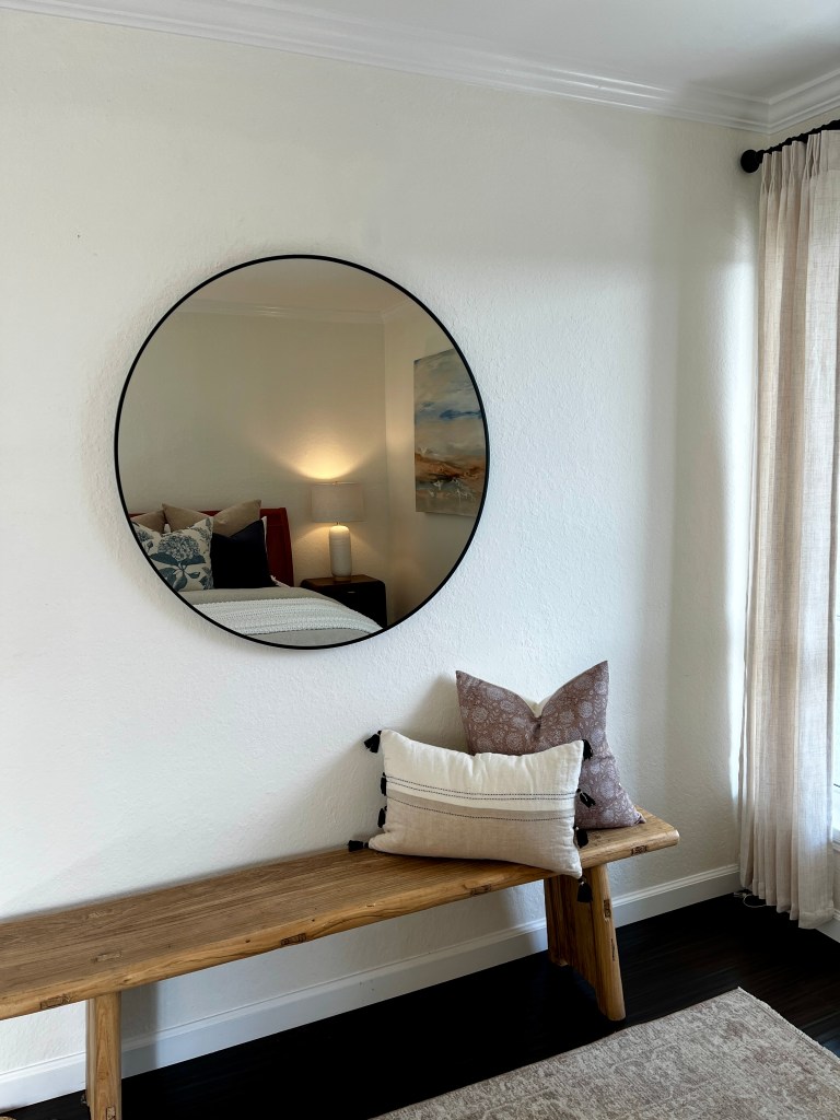A round black-framed mirror hangs on a wall above a wooden bench adorned with decorative pillows, including a patterned pillow and a plain one, creating a cozy interior setting.