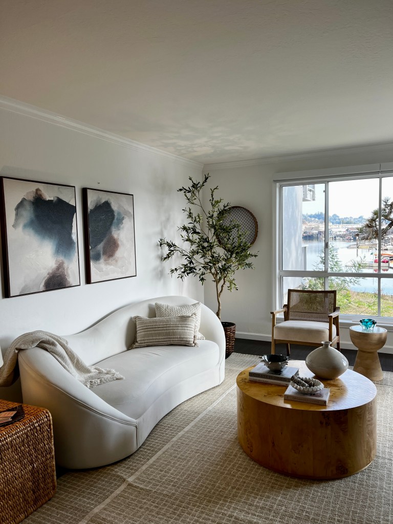 Modern living room featuring a white curved sofa, a round wooden coffee table, and a woven accent chair. The space includes abstract wall art, a decorative plant, and a large window with a view of the outdoors.