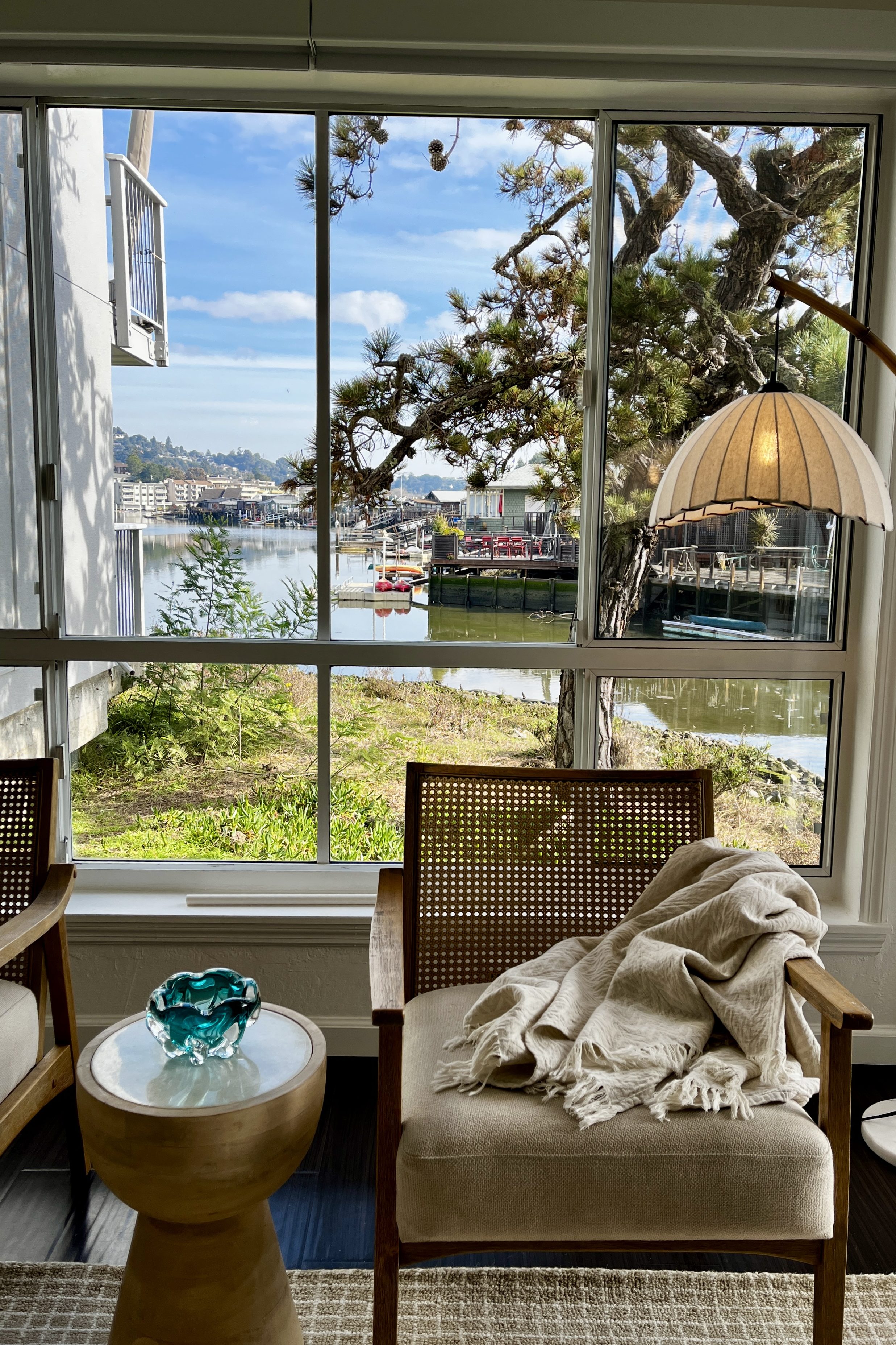 A cozy interior scene featuring two wooden chairs with cushions, a round side table with a decorative glass bowl, and a soft blanket draped over one chair. The window offers a view of a tranquil waterfront with boats and lush greenery, under a partly cloudy sky.