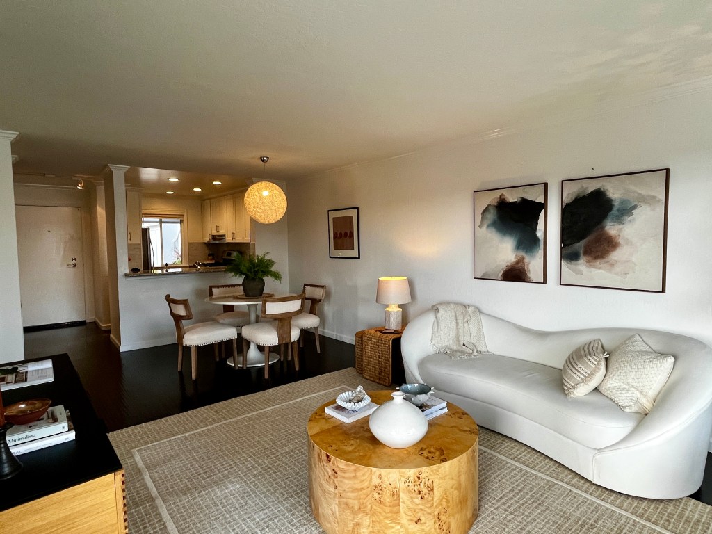 Modern living room featuring a white curved sofa, a round wooden coffee table, and decorative items. A dining area with four chairs is visible in the background, along with a well-lit kitchen. Wall art adds a contemporary touch to the space.